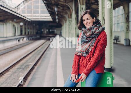 Die freudig junge Europäische Frau trägt roten Pullover, Schal, hält Handy, sitzt in der Tasche, reist gern, posiert am Bahnhof, lächelt glücklich. Peopl Stockfoto