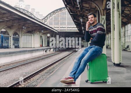 Lifestyle-Konzept für jeden Tag. Gutaussehender Mann trägt Pullover und Jeans, posiert auf Bahnsteigplattform, lehnt sich an Tasche, hat Telefongespräche, fokussiert in Stockfoto