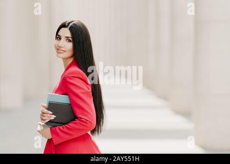 Seitlicher Schuss von eleganter Brunette-Dame mit dunkellangem geradem Haar, in modischem roten Anzug gekleidet, hat Maniküre, trägt Tagebuch, steht draußen Stockfoto