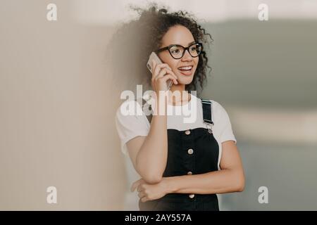 Lächelnde junge, lockige Frau mit einem sanften Lächeln, spricht auf dem Handy, hat ein freundschaftliches Gespräch, trägt ein legere weißes T-Shirt und Overalls, die beiseite gerichtet sind. Stockfoto