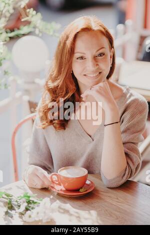 Menschen- und Freizeitkonzept. Positive, rothaarige Frau mit einem sanften Lächeln hält die Hand unter dem Kinn, mit legeren Pullover bekleidet, und trinkt aromatischen Kaffee Stockfoto