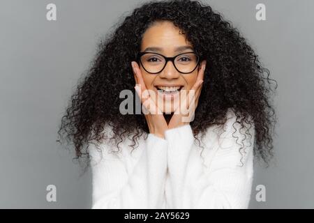 Nahaufnahme der entzückten afroamerikanischen Frau berührt beide Wangen mit Händen, drückt positive Emotionen aus, trägt transparente Brille und weiße sw Stockfoto