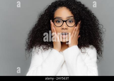 Close up Portrait von schockiert betroffene junge Frau mit dunkler Haut und lockiges Haar, hört erschreckende Nachrichten, hält die Hände auf die Wangen, trägt eine Brille und w Stockfoto