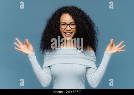 Positive Afrikanische amerikanische Dame macht okay Geste mit beiden Händen, meditiert, Indoor, trägt modische Pullover und eyewear, gut gelaunt, isolat Stockfoto