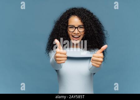 Fröhliche schöne Frau mit buschigen Afro-Haaren gibt den Daumen nach oben, billigt eine schöne Idee, trägt einen modischen Pullover, isoliert über blauem Hintergrund. Sein Fein, ich agre Stockfoto