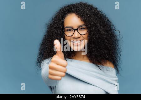 Portrait von dunkelhäutige Frau hat Daumen hoch, zeigt Zustimmung oder Genehmigung, hat ein Lächeln auf dem Gesicht, trägt eine Brille und Jumper, Gesten innen gegen Blu Stockfoto