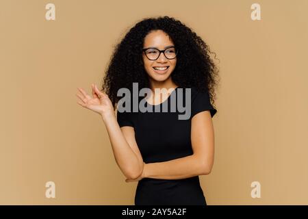 Lächelnde Frau mit Afro Haarschnitt, macht okay Geste, sieht glücklich beiseite, stimmt mit Mäusevorschlag überein, trägt optische Brille und legeren schwarzen T-Schir Stockfoto