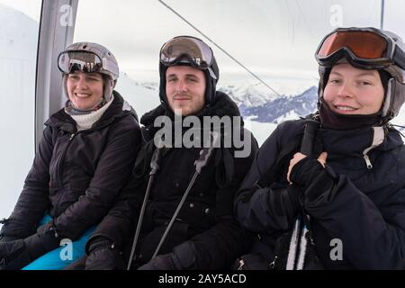 Junge Erwachsene sitzen in einer Seilbahn, die auf einem Hang verläuft, tragen Helm und Schutzbrille und halten an einem bewölkten Tag Stöcke mit Schneeberg im Hintergrund Stockfoto