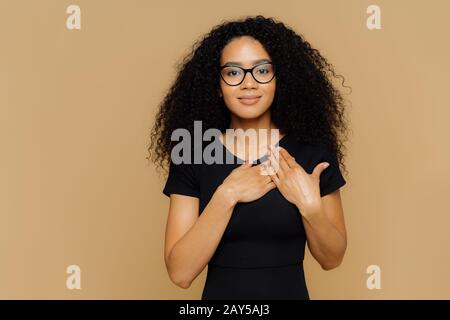 Schöne dunkelhäutige Frau hält Handflächen in der Nähe zu hören, drückt Dankbarkeit, Dankbarkeit aus, hat freundlichen Gesichtsausdruck, trägt Brille und lässige Bla Stockfoto