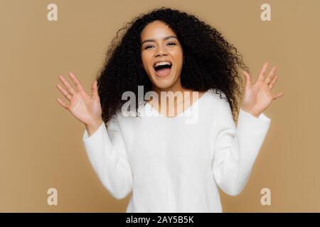 Emotional froh afroamerikanische Frau mit knackigen schwarzen Haaren, hebt Hände, Gesten aktiv, beobachtet etwas wunderbares und tolles, trägt weiches Whi Stockfoto