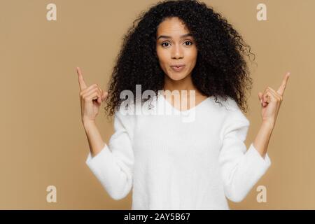 Foto der selbstbewussten Frau hat buschige Afro-Frisur, zeigt beide Zeigefinger oben, bekleidet mit weißem Pullover, steht vor braunem Hintergrund, Advertis Stockfoto