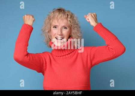 Fröhliche Seniorin im roten Pullover mit den Fäusten, die ihren Erfolg feiern. Positive menschliche Emotionen im Gesicht. Studio gedreht Stockfoto