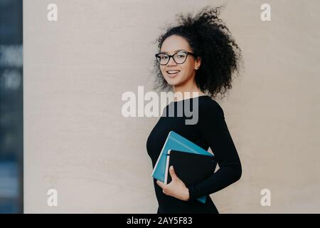 Seitlicher Schuss von schönen dunkelhäutigen Studenten der geschweiften Hochschule, gekleidet in schwarzem Casual Outfit, trägt Lehrbuch, Notizblock, lächelt positiv, isoliert o Stockfoto