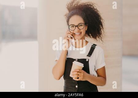 Angenehm aussehendes lockiges Teenager-Mädchen trägt transparente Brille, spricht auf dem Handy, hat Kaffeepause, gekleidet mit weißem T-Shirt und Overalls, genießt Stockfoto