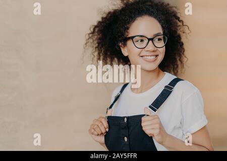 Foto des freudigen afro-amerikanischen Teenagers mit lockigen Haaren, lächelt breit, blickt weg, trägt transparente Brille und Overalls, hat gute Laune, verbringt f Stockfoto