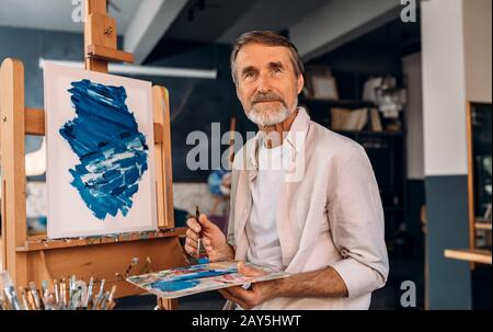Porträt eines älteren Malers, der mit einer Pinsel Palette hält und wegschaut. Künstler in seinem Studio. Stockfoto