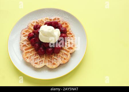 Köstliche Waffeln mit heißen Kirschen und Creme auf einem weißen Teller, gelber Hintergrund mit Platz für Kopien Stockfoto