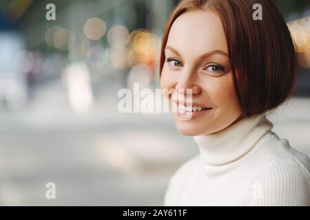 Seitlicher Schuss von angenehm aussehender Frau mit Make-up, zartes Lächeln, bekleidet mit weißem Turnleneck-Pullover, steht vor verschwommenem Hintergrund mit Copy s Stockfoto