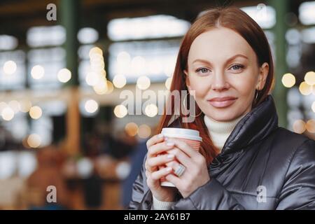 Außenaufnahme des attraktiven weiblichen Modells mit gesunder Haut, braunem Haar, hält Kaffee zum Mitnehmen, genießt einen guten Drink, in der Jacke gekleidet, wartet fo Stockfoto