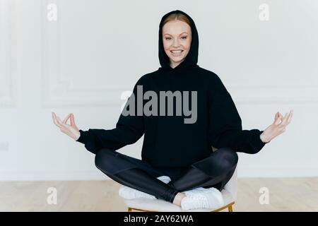 Horizontaler Schuss optimistischer Frauen übt Yoga, meditiert auf dem Stuhl, sitzt in lotus-pose, trägt schwarzes Sweatshirt, weiße Sportschuhe, hat einen frohen Ausdruck Stockfoto