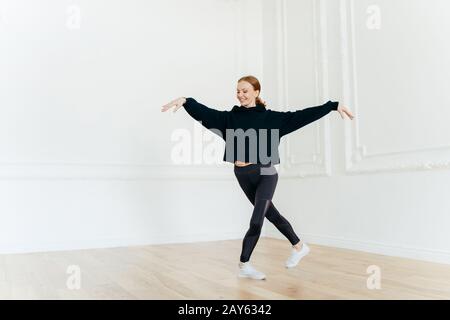 Professionelle Ballerina übt Tanz im Saal, streckt Hände, steht mit gekreuzten Beinen, hat ein sanftes Lächeln im Gesicht, bekleidet in schwarzem Sportswea Stockfoto