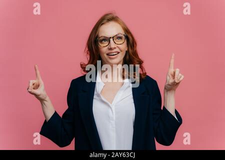 Foto der freudigen rothaarigen Frau blickt nach oben, zeigt die Finger nach oben, hat zufriedenen Gesichtsausdruck, zeigt etwas, trägt Brille, f. Stockfoto