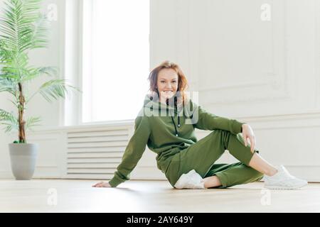 Fröhliche Fitness rothaarige Frau lehnt sich mit der Hand auf dem Boden, hat Zeit für Erholung nach gymnastischen Übungen, trägt grünen Trainingsanzug, posiert innen, hat Athleti Stockfoto