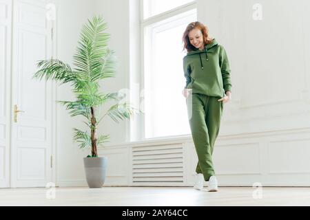 Die lange Aufnahme einer aktiven, freudigen jungen Frau mit schlankem Körper hält die Hände in den Taschen des Trainingsanzugs, konzentriert nach unten, bereit für Herz-Kreislauf-Übungen, ha Stockfoto