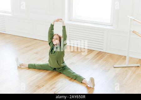 Starke flexible, geschweifte, rothaarige Frau spaltet das Seitenbein, hebt die Hände, will einen perfekten Körper haben, trägt Sportkleidung, hat eine Ausbildung im Ballettgestühl Stockfoto