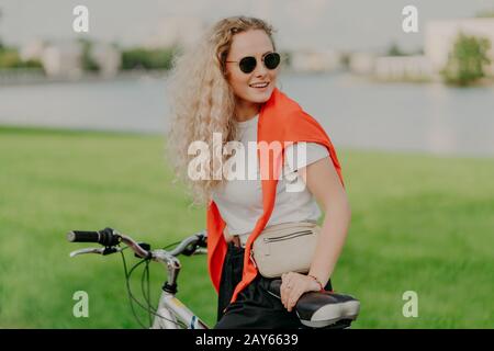 Foto der attraktiven, lockig behaarten Frau, die auf der Seite fokussiert ist, trägt Schattierungen, gekleidet in lässiger Kleidung, posiert auf dem Fahrrad, wartet auf Freundinnen, die in die Ferne blicken Stockfoto