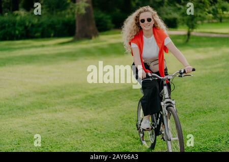 Menschen, Natur, Ruhe, Lifestyle-Konzept. Fröhliche lockige Frau fährt mit dem Fahrrad zwischen grünem Gras, bewegt sich aktiv, will fit sein, erkundet neue Orte in Co Stockfoto