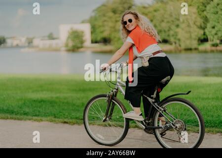 Bild von weiblichen Modelfahrten Fahrrad, sieht mit fröhlichem Ausdruck beiseite, trägt Sonnenbrille, atmet frische Luft, posiert in der Nähe von See und grünen Bäumen, Bucht Stockfoto