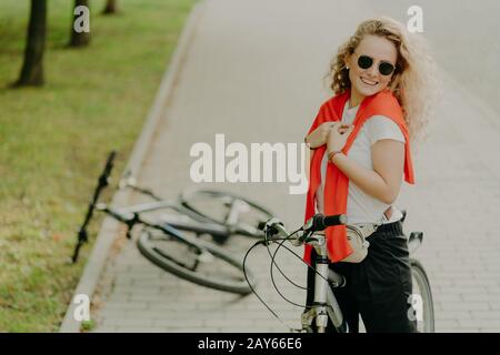 Fröhliche, lockig behaarte Weibchen hält unterwegs an, posiert auf dem Fahrrad auf der Straße, Friends Bike liegt in der Nähe, trägt Sonnenbrille, T-Shirt und Hose, trägt Littl Stockfoto