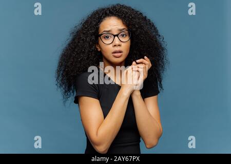 Foto von peinlich angeprangerter afro-amerikanischer Frau hält die Hände zusammen, sieht nervös auf die Kamera, hat einige Probleme, trägt legere schwarze T-Shirts, steht wieder Stockfoto