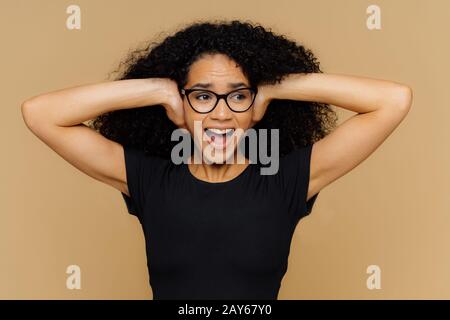 Bild der emotionalen afroamerikanischen Frau bedeckt Ohren, schreit laut an, kann keine Geräuschanforderungen stehenlassen Musik ausschalten, gekleidet in lässigem schwarzem T-Shirt, optisch Stockfoto