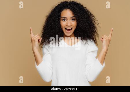 Konzept für Menschen, Werbung und Emotionen. Froh junge Frau zeigt mit Zeigefingern nach oben, zeigt einen tollen Artikel im Verkauf, trägt einen weißen Pullover, exkl. Stockfoto