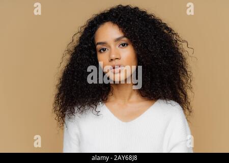 Das Porträt eines ernsthaften, dunkelhäutigen Weibchens mit flauschigen schwarzen Haaren hat ein minimales Makeup, wirkt ruhig auf die Kamera, trägt einen weißen Pullover, steht wieder Stockfoto