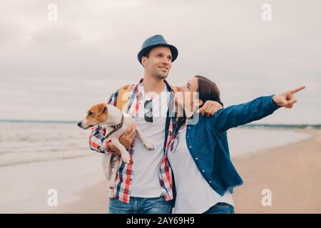 Horizontaler Schuss von liebendem Paar umarmen sich und gehen draußen auf dem sandigen Strand, tragen Lieblingshund, schöne Frau zeigt mit Zeigefinger in die Ferne, Stockfoto