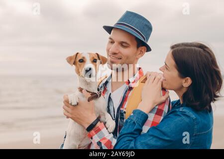 Positive weibliche und männliche Touristen stehen dicht über der schönen Meereslandschaft, schauen sich ihren Hund an, genießen Urlaub an der Küste, tragen modischen Outf Stockfoto