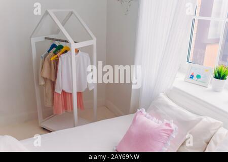 Gemütliches, geräumiges helles Zimmer mit modischer Kleidung an Kleiderbügeln, bequemem Bett mit weißem Schlafzimmer und weißen Wänden. Häusliche Atmosphäre Stockfoto