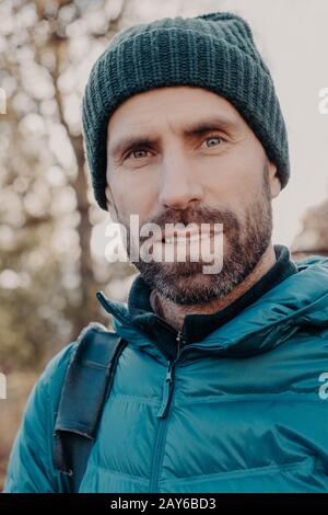 Nahaufnahme des blauäugigen Mannes hat dunkle Borsten, schaut ernsthaft auf die Kamera, trägt Hut und Jacke, stellt sich draußen, spaziert im Freien, hat angenehmen Appetit Stockfoto