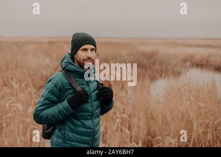 Penibel unshaven Kerl trägt Hut, grüne Jacke und Handschuhe, trägt Rucksack, posiert vor gelbem Herbstfeldhintergrund, ist draußen spazieren, atmet Stockfoto