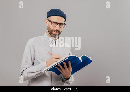 Horizontale konzentrierter männlichen Texter schreibt Ideen für neue Strategie, hält blau Notepad, trägt eine Brille für gute Sicht, hält Bleistift, sta Stockfoto