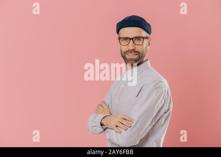 Studio geschossen von Selbstsicher bärtiger Mann mit blauen Augen, trägt stylischen Hut und weissen Hemd, hält die Arme verschränkt, Modelle über rosa Hintergrund mit kostenlosen sp Stockfoto