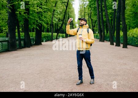 Außenporträt junger, gutaussehender Tourist in stilvoller Kleidung, die Rucksack hält und Kaffee in die Kamera seiner Smartphone-Herstellung wirft Stockfoto