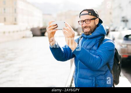 Stilvoller Mann mit blauen, bezaubernden Augen und Bart mit blauem Anorak, Brille und Kappe macht selfie mit Handy vor Großstadthintergrund. Jung Stockfoto