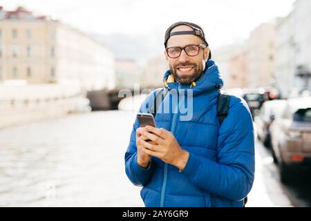 Glücklicher Mann mit Bart mit Brillen, die in blauem Anorak-Haltersack gekleidet sind, und Mobilgerät, der einen fröhlichen Look hat, der in der Stadt unterwegs ist und Sehenswürdigkeiten beobachtet Stockfoto