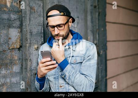 Ernster gebärter bärtiger Mann in Kappe und Denimjacke, der gegen eine gerissene Wand steht, die Smartphone-Pfosten hält und sie über das soziale Netz teilt Stockfoto