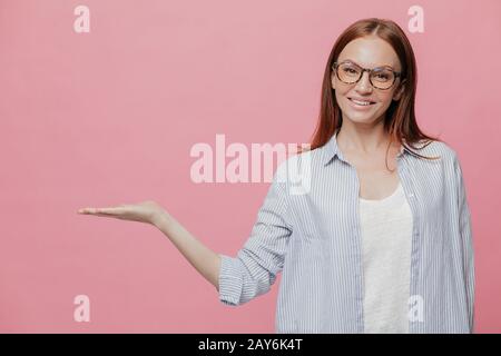 Studio shot zufrieden kaukasische Frau hält Palm erhoben, trägt optische Gläser, zeigt etwas über Kopieren, enthält Leerzeichen, hat pleasa Stockfoto