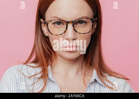 Nahaufnahme des hübschen weiblichen Modells trägt eine große optische Brille, hat braune Haare, wirkt ernsthaft mit selbstbewusster Ausdrucksweise, isoliert über rosa Backgrou Stockfoto